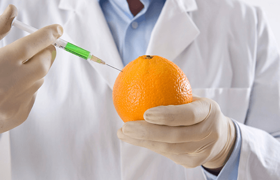 Photo of orange being held by someone is a lab coat with rubber gloves on injecting the orange with unknown green substance in a syringe
